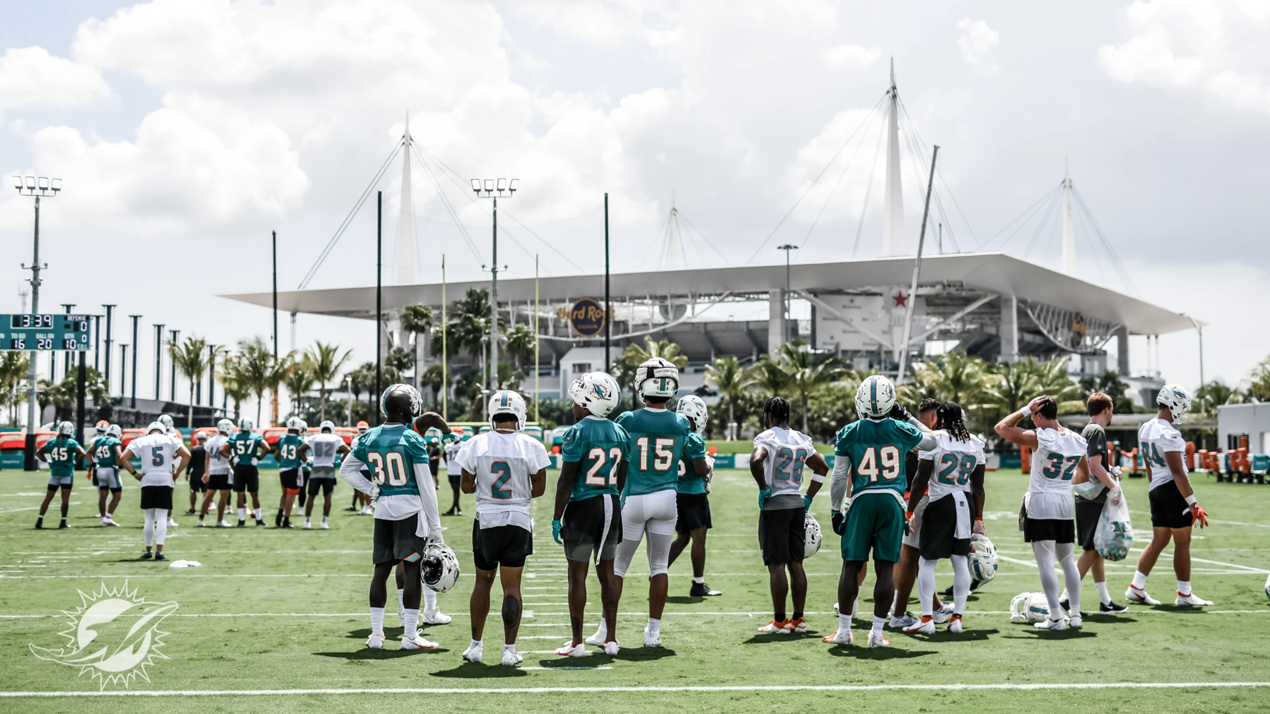 Miami Dolphins players practicing at Baptist Health Training Complex in Miami Gardens with fans watching from grandstands, palm trees and modern facility visible in background