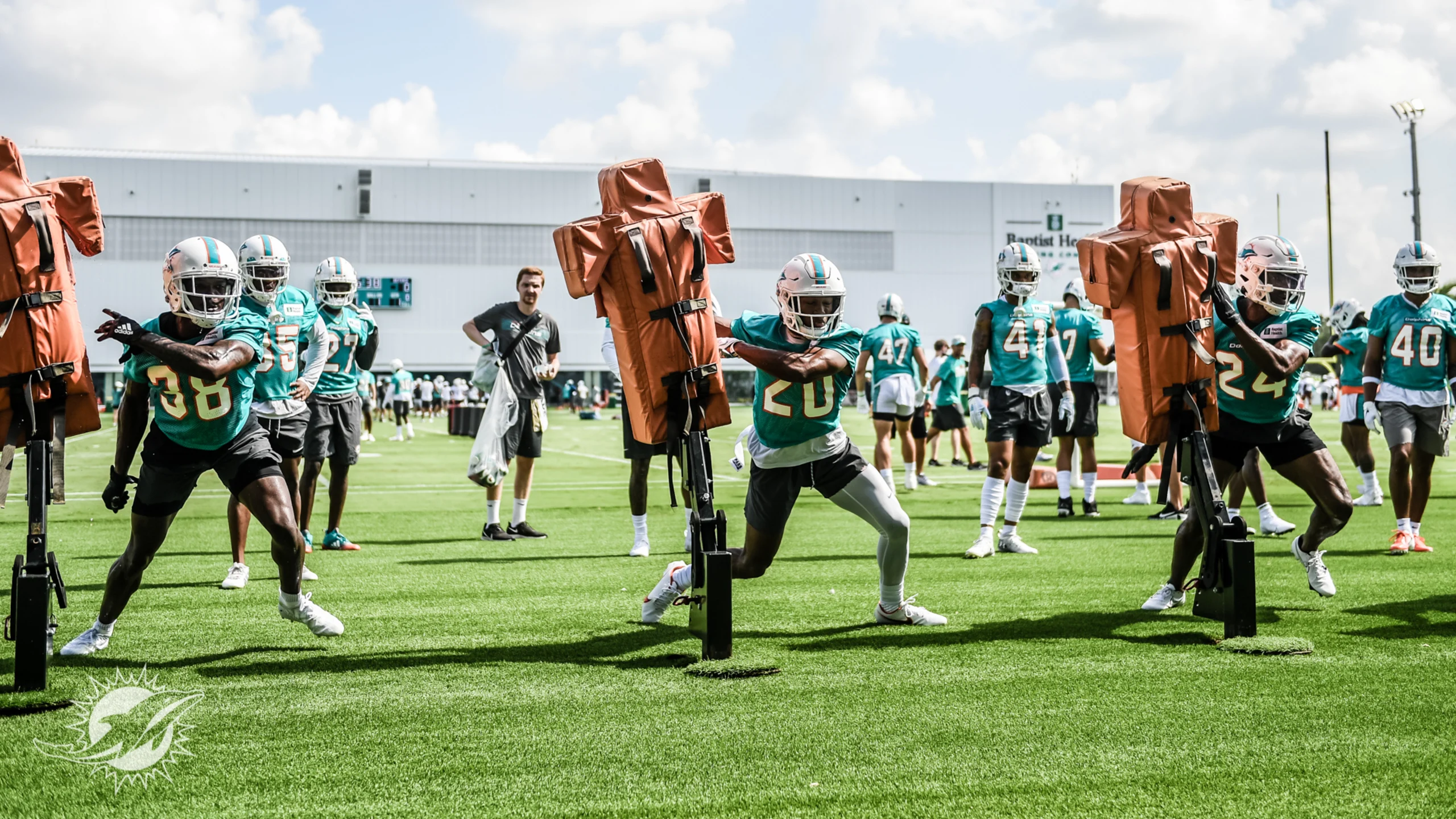 Miami Dolphins players running drills on practice field at Baptist Health Training Complex with coaches observing and fans in background